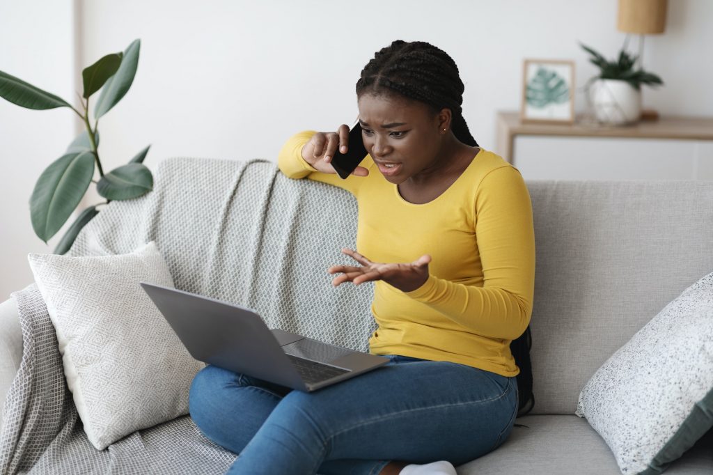Frustrated black lady using laptop and talking on cellphone at home